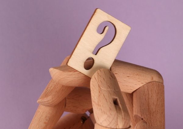 Wooden mannequin hand holding a carved question mark against a purple background.